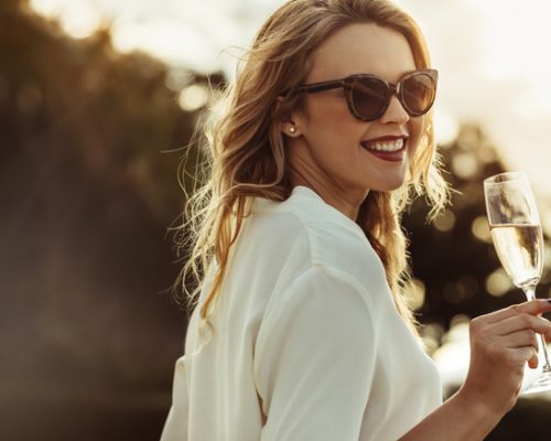 Elegant woman in sunglasses with a glass of wine outdoors. Smiling caucasian female having wine and looking backwards.